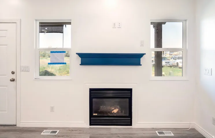 Bright living space featuring a wood-burning fireplace with black surround and hearth, two large windows with blue window treatments, neutral wall finishes, and wood-finish flooring throughout