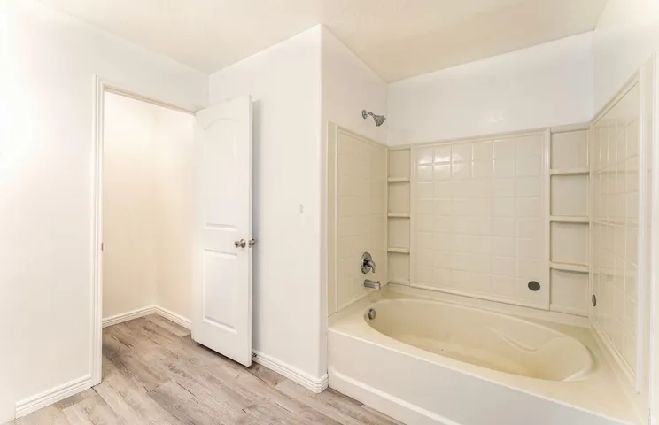 Spacious primary bathroom featuring a soaking tub with integrated surround, ceramic tile walls, recessed shelving, wood-finish flooring, and a separate water closet with door
