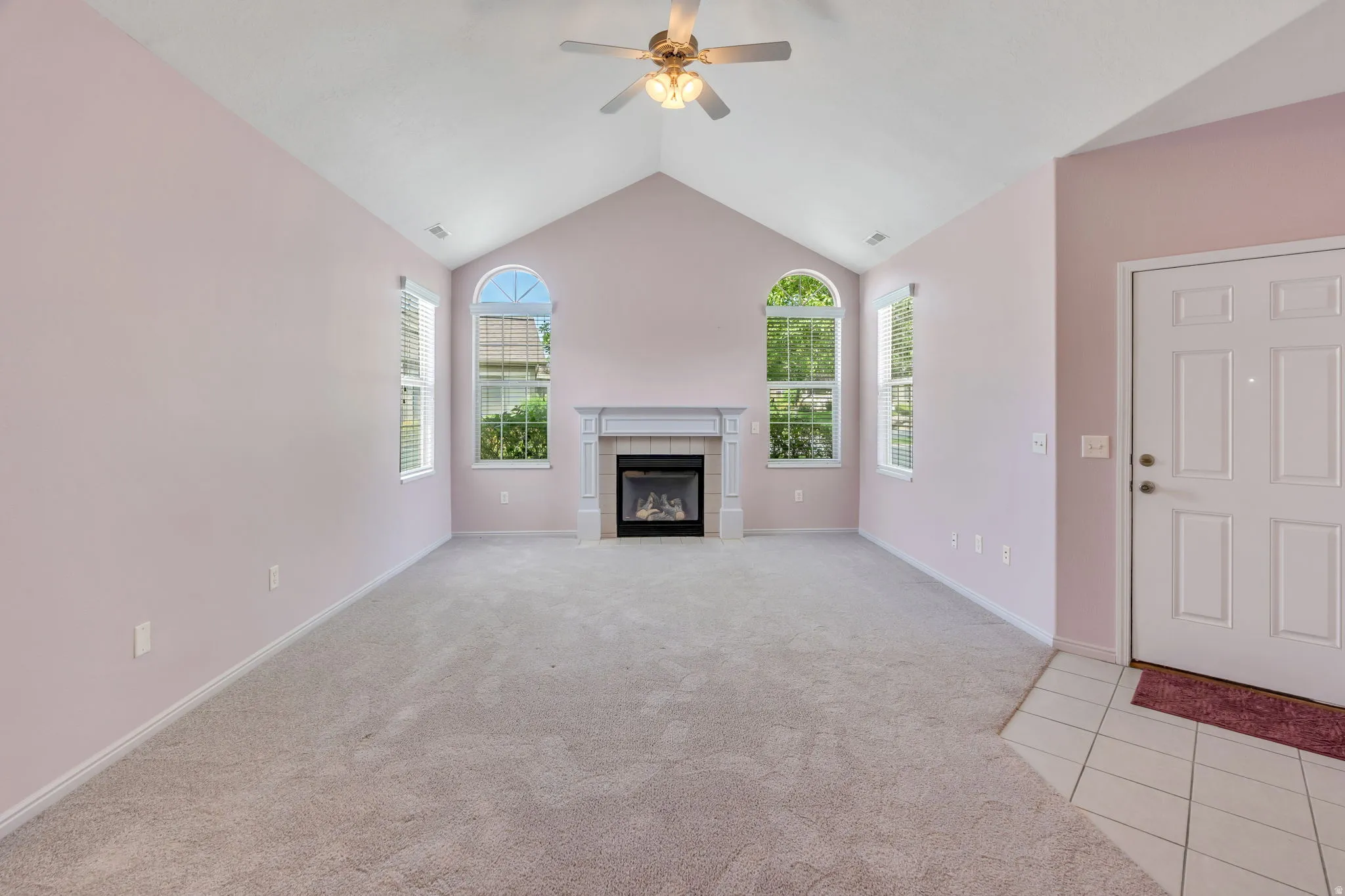 Vaulted ceiling living space featuring a fireplace with white mantel, two arched-top windows, ceiling fan, and light gray wall-to-wall carpeting