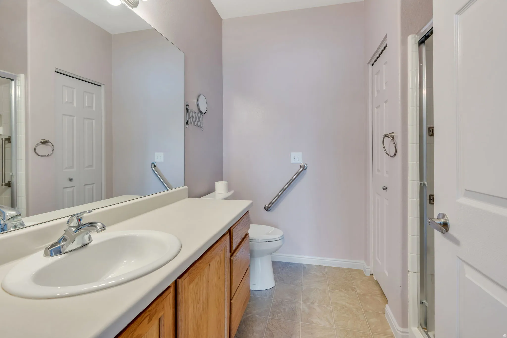 Bathroom featuring a single vanity with a white countertop and wood-finish cabinetry, a large wall-mounted mirror, a commode with a grab bar, and a glass-enclosed shower