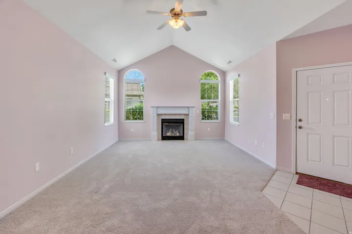 Vaulted ceiling living space featuring a fireplace with white mantel, two arched-top windows, ceiling fan, and light gray wall-to-wall carpeting