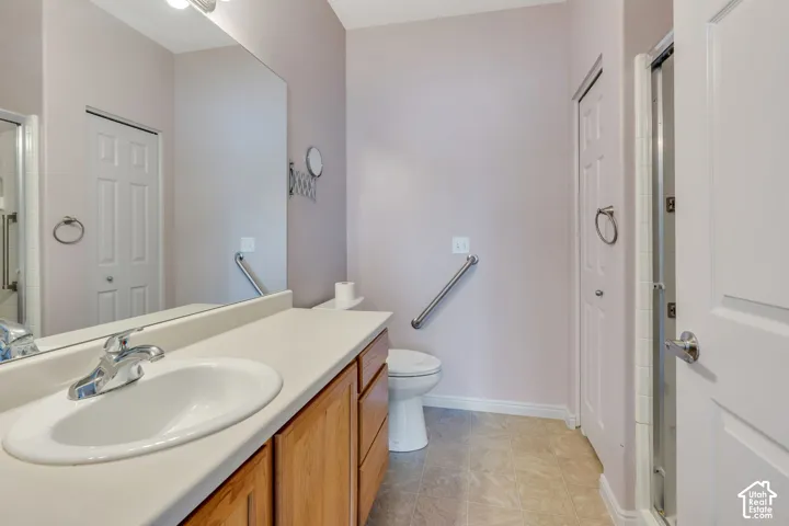 Bathroom featuring a single vanity with a white countertop and wood-finish cabinetry, a large wall-mounted mirror, a commode with a grab bar, and a glass-enclosed shower