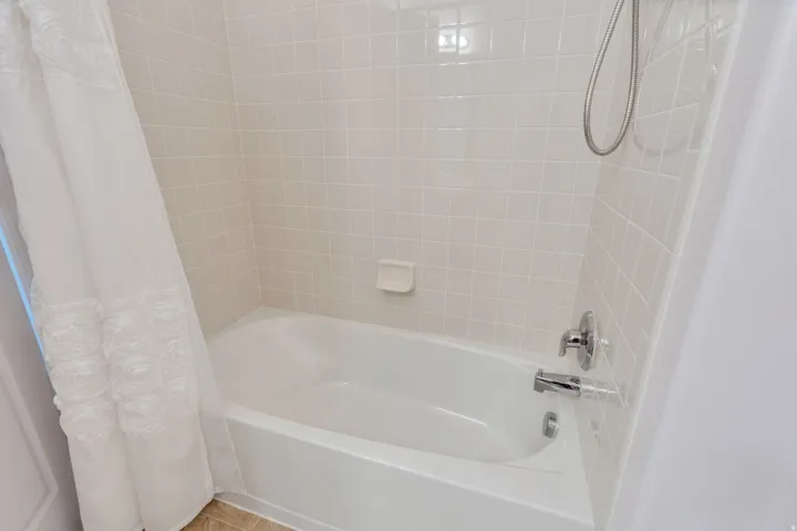 White tub and shower combination with light-toned square tile surround, chrome fixtures, and a shower wand