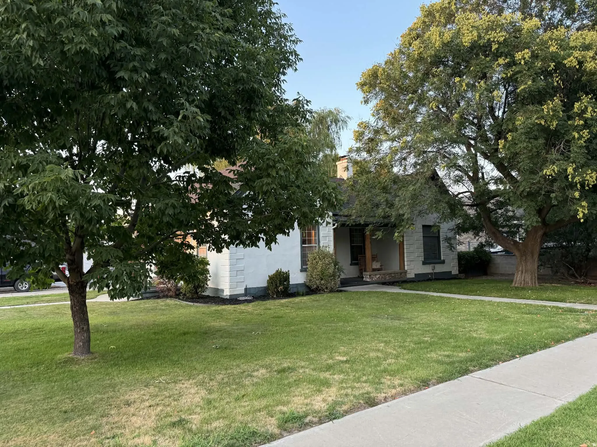 White facade with dark trim, featuring a covered porch, established lawn, mature trees, and a concrete sidewalk