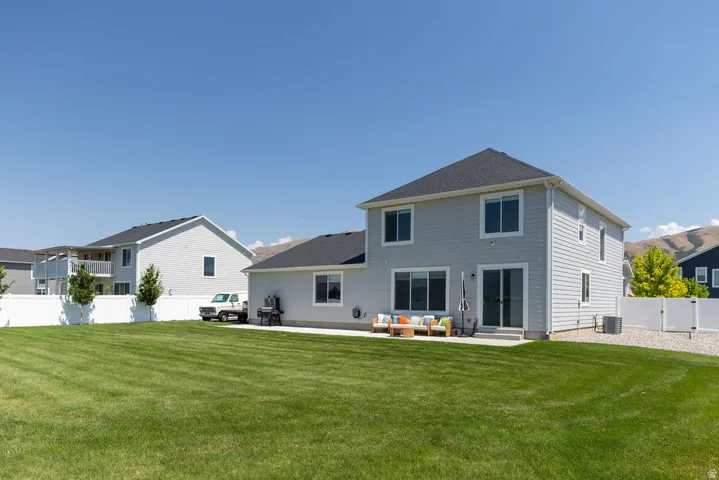 Expansive grass lawn with a concrete patio, light gray siding, multiple windows, and a white privacy fence