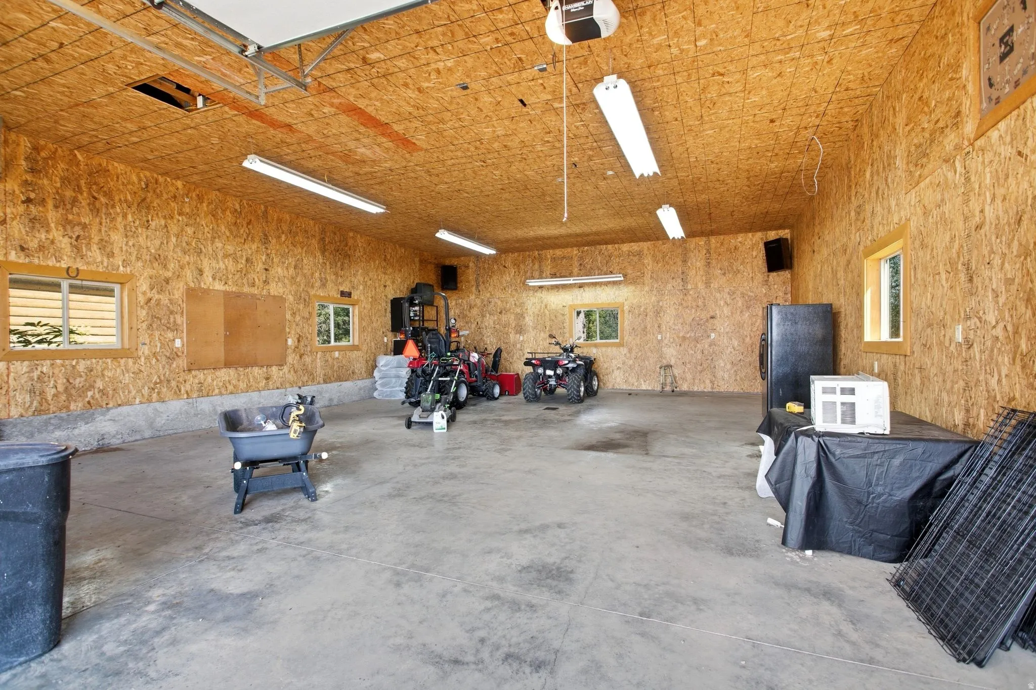 Spacious garage featuring a concrete floor, exposed OSB walls and ceiling, multiple windows, and fluorescent overhead lighting
