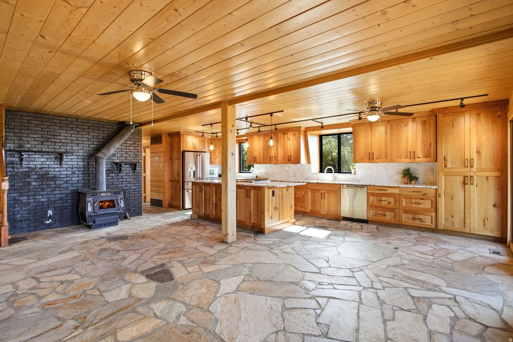 Open-concept living space with flagstone flooring, wood plank ceilings, and a black brick fireplace featuring a wood-burning stove