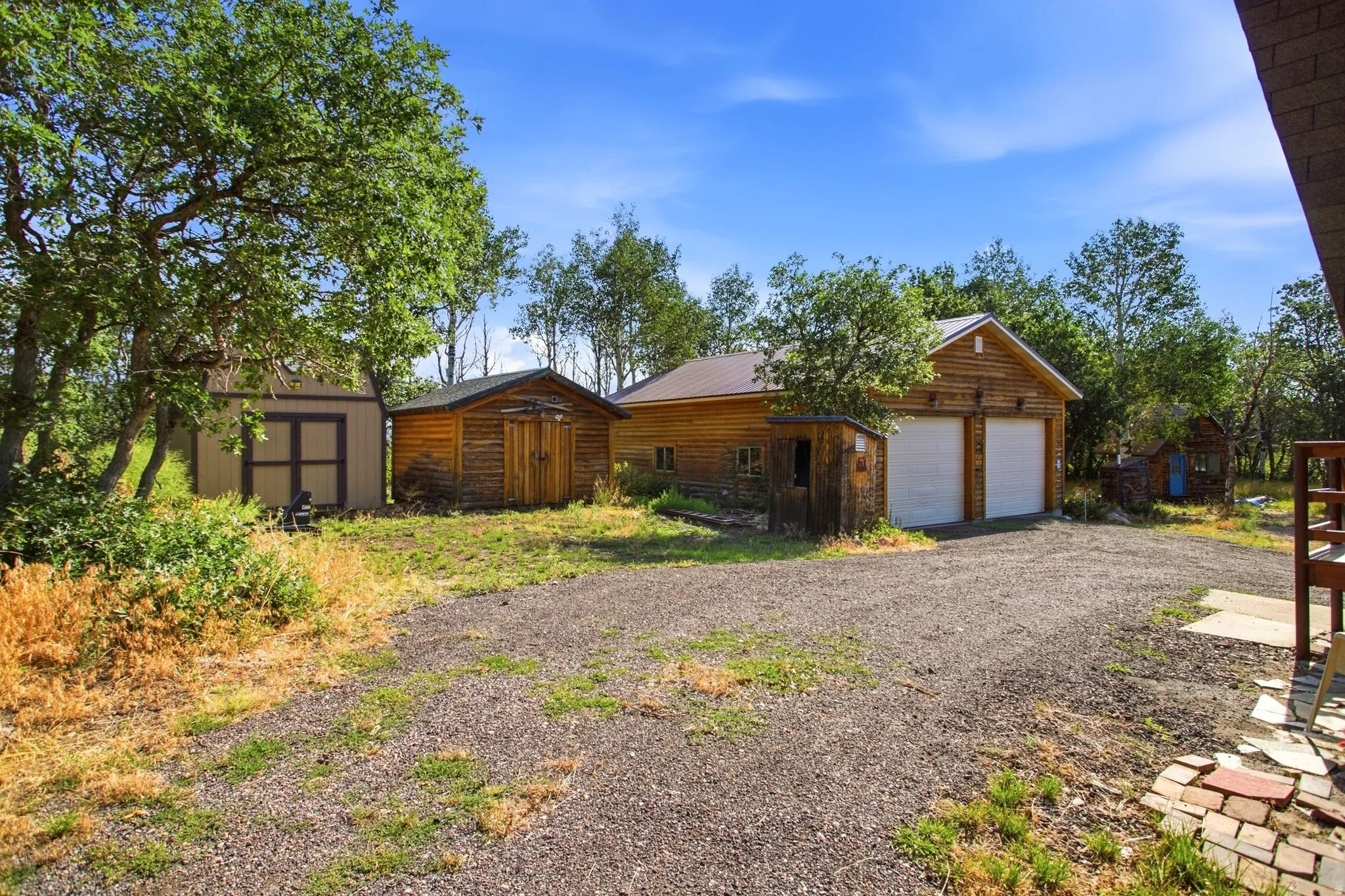 Gravel driveway with a two-car garage featuring a log-style exterior, two sheds, and surrounding mature trees