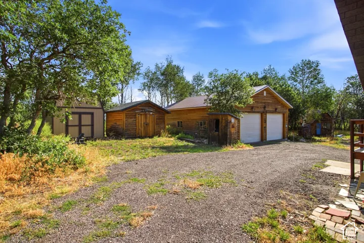 Gravel driveway with a two-car garage featuring a log-style exterior, two sheds, and surrounding mature trees