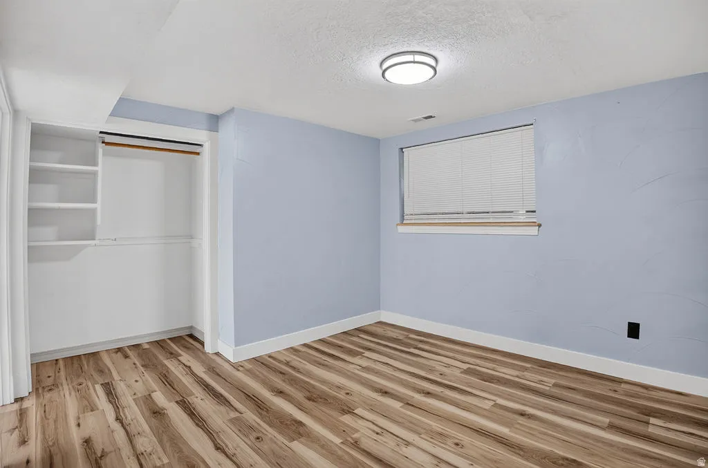Room featuring wood-finish flooring, light blue walls, white trim, a built-in closet with shelving and hanging rods, a window with blinds, and a flush-mount ceiling light fixture