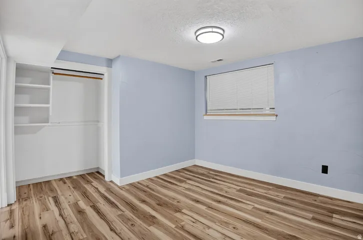 Room featuring wood-finish flooring, light blue walls, white trim, a built-in closet with shelving and hanging rods, a window with blinds, and a flush-mount ceiling light fixture