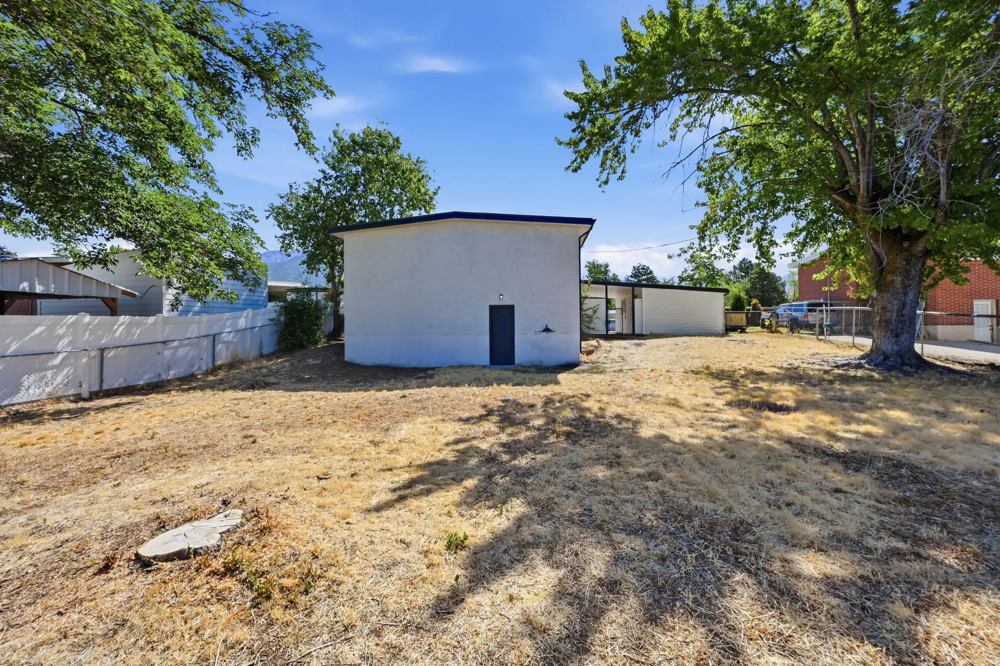 Expansive outdoor space featuring a white stucco structure with a dark blue entry door, surrounded by a chain-link fence and mature trees