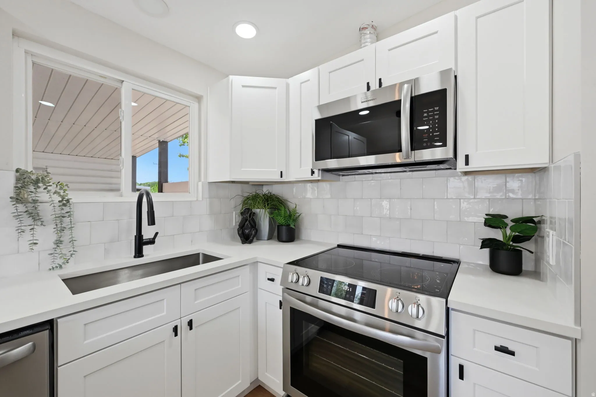 Contemporary kitchen featuring white shaker cabinetry, stainless steel appliances, a black matte pull-down faucet, a farmhouse-style sink, and light-colored countertops