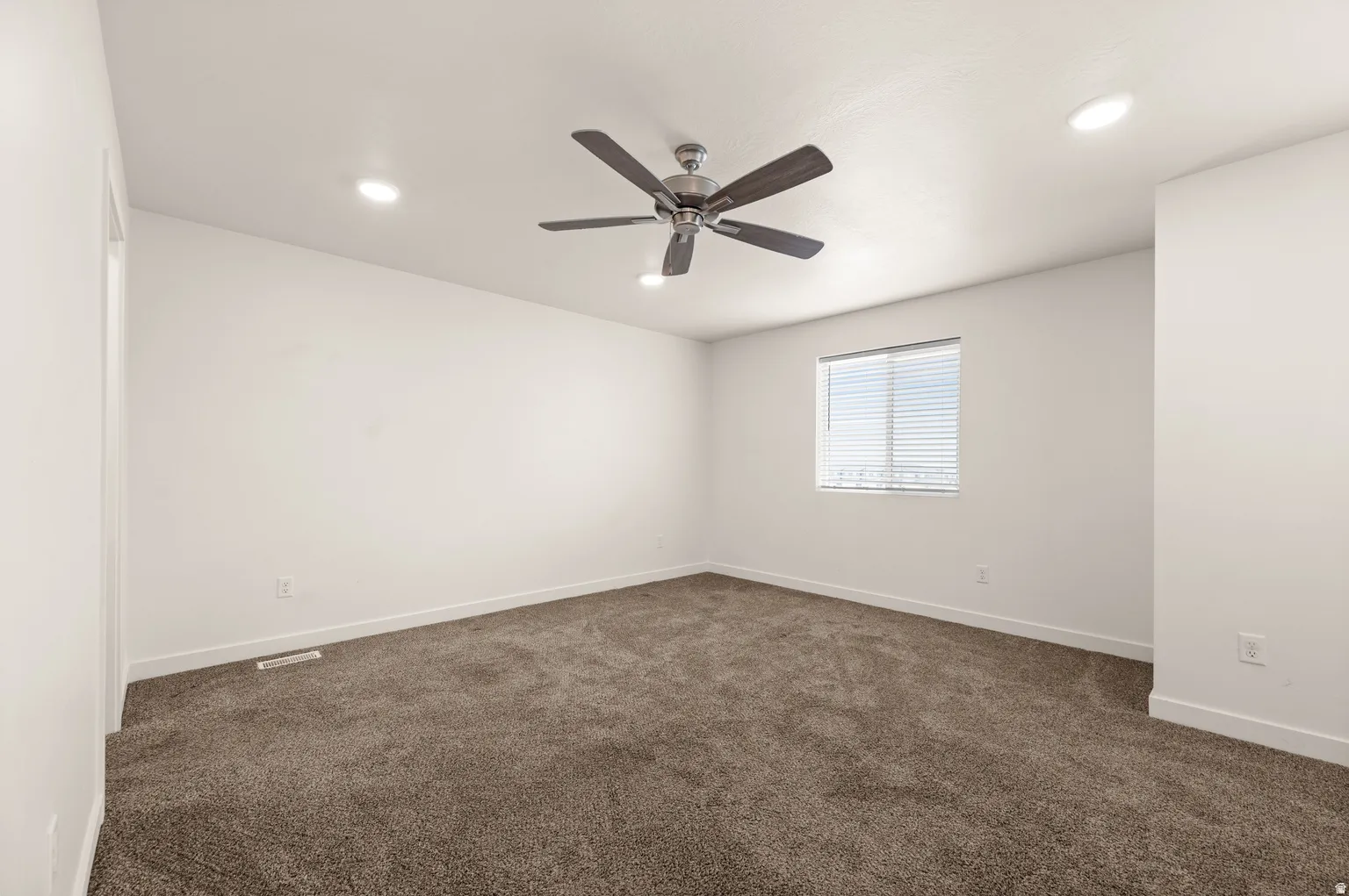 Spacious room featuring neutral carpeting, recessed lighting, a ceiling fan, and a single window with blinds