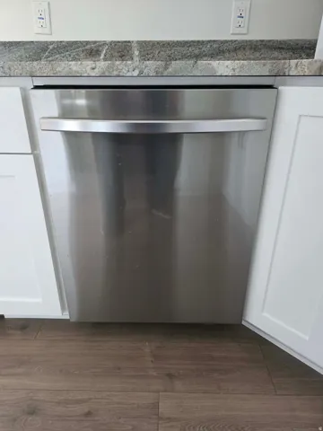 Stainless steel dishwasher with a bar handle, set within white cabinetry and under a speckled stone countertop