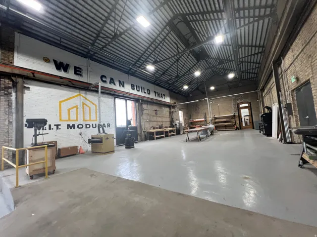 Spacious industrial interior featuring a high vaulted ceiling with exposed metal trusses, brick walls, and a light gray concrete floor