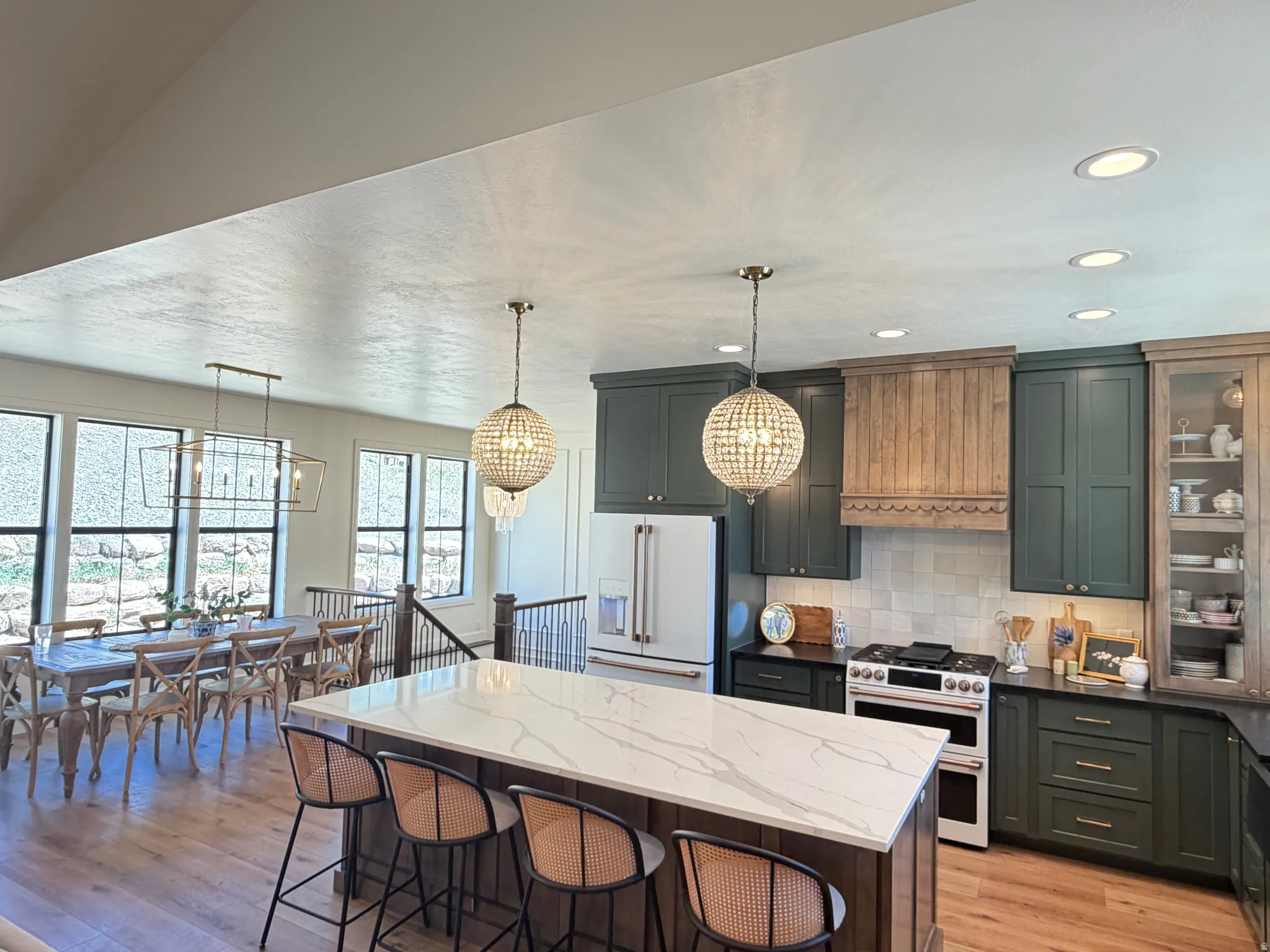 Open-concept kitchen featuring an oversized island with seating, dark green cabinetry, a wood-finish range hood, and light wood-finish flooring