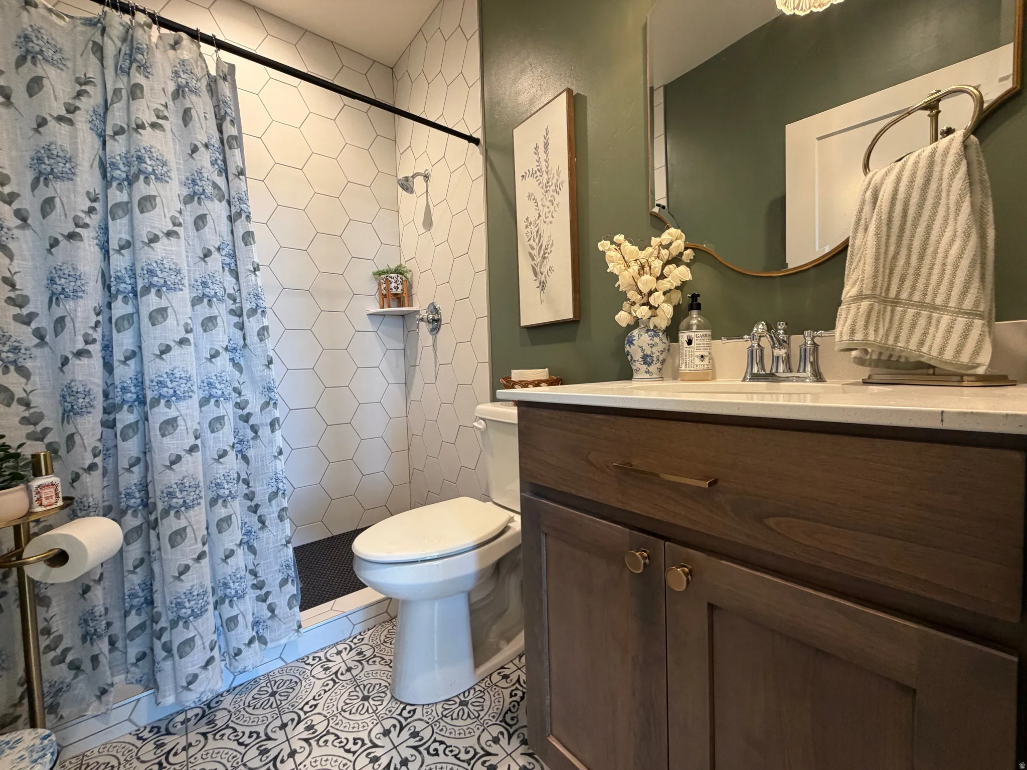 Hall bathroom featuring patterned floor tiling, white hexagonal wall tiling, a built-in shower, a wood-finish vanity with a white countertop, and a circular wall mirror