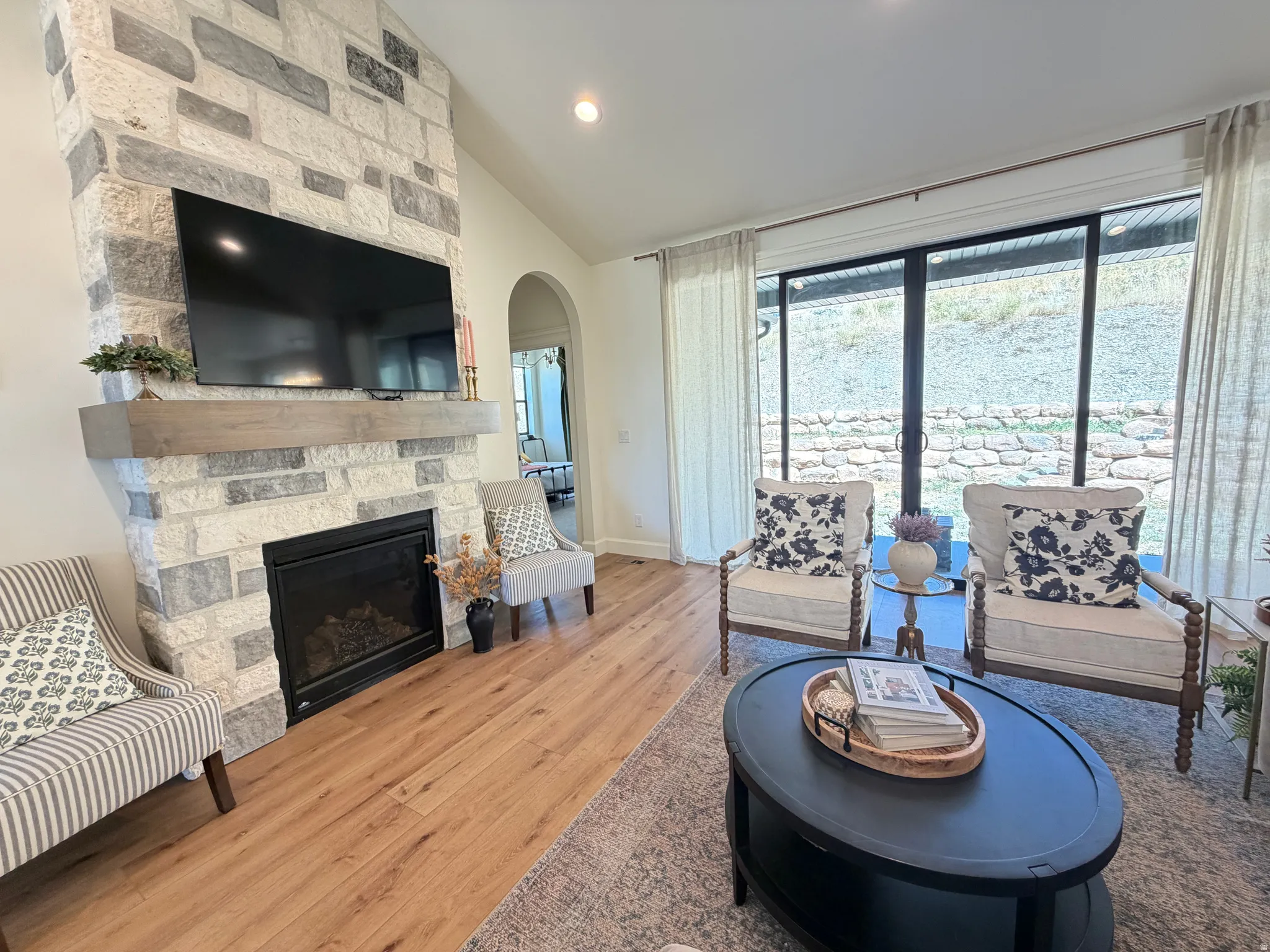 Vaulted living area featuring a floor-to-ceiling stone fireplace with a wood-finish mantel, wood-finish flooring, and large glass sliding doors providing ample natural light