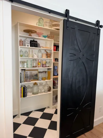 Custom built-in pantry featuring extensive shelving, a black and white checkerboard floor, and a decorative black sliding barn door with dark hardware