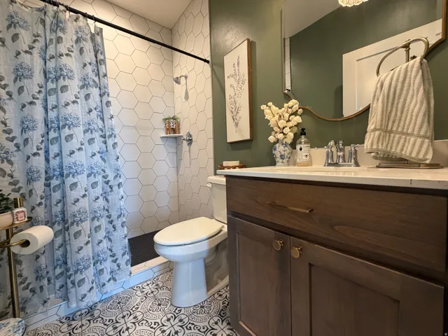 Hall bathroom featuring patterned floor tiling, white hexagonal wall tiling, a built-in shower, a wood-finish vanity with a white countertop, and a circular wall mirror