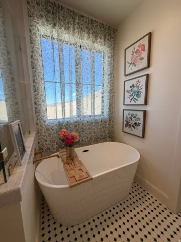Freestanding soaking tub with a wall-mounted faucet, complemented by patterned sheer window treatments and hexagon tile flooring