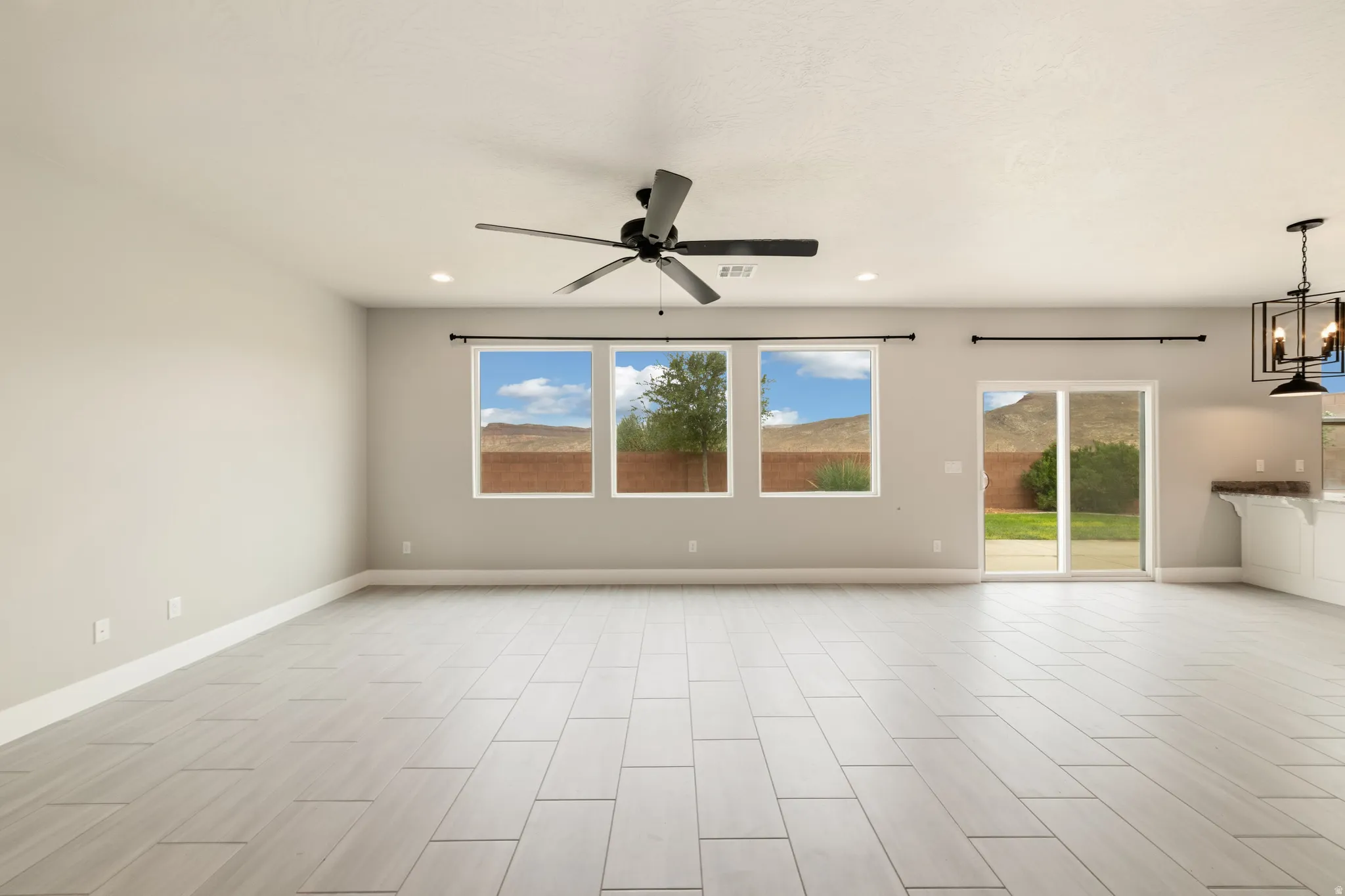 Spacious living area featuring light-toned tile flooring, three large windows, a ceiling fan with integrated lighting, and a sliding glass door leading to the exterior