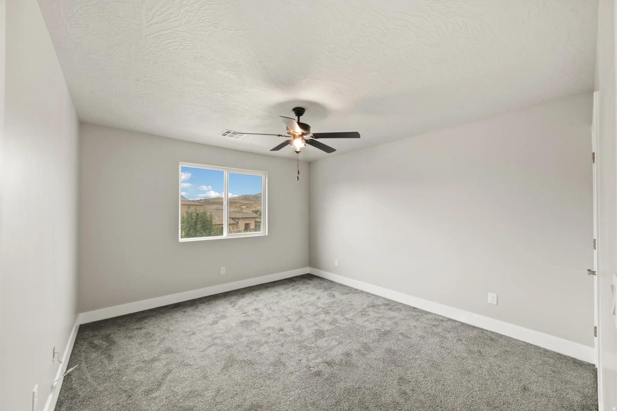 Spacious interior room featuring a large window, neutral wall tones, gray carpeting, a ceiling fan with light, and white trim