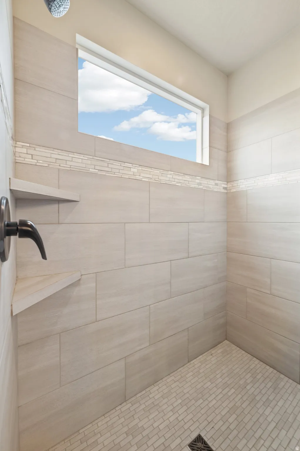 Tiled shower enclosure featuring a rectangular transom window, a dark bronze showerhead, and a built-in corner shelf