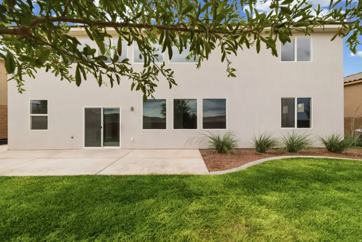 Rear exterior featuring a concrete patio, sliding glass door, multiple windows, manicured lawn, and planted landscape bed with decorative rock