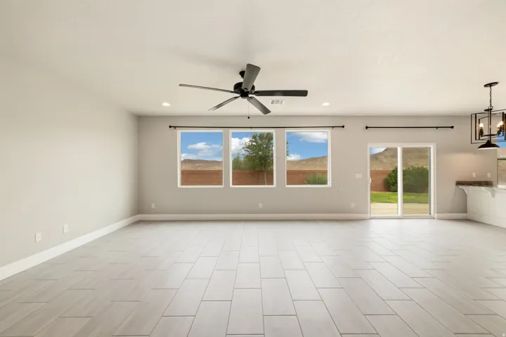 Spacious living area featuring light-toned tile flooring, three large windows, a ceiling fan with integrated lighting, and a sliding glass door leading to the exterior