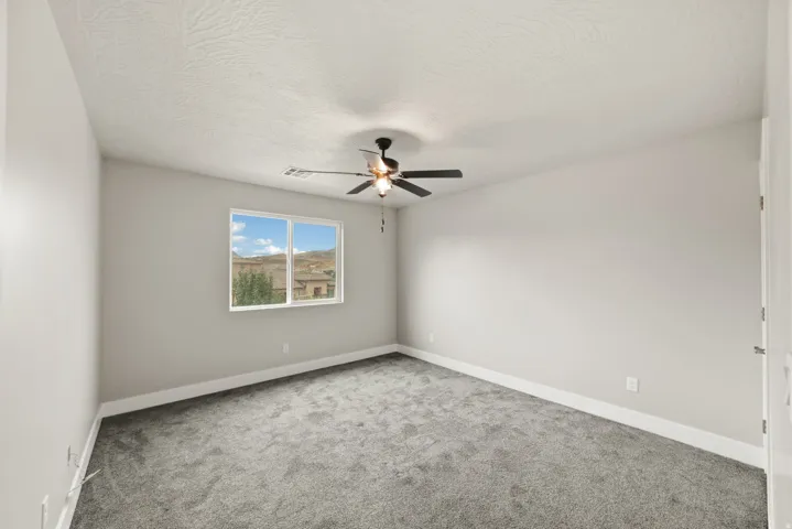Spacious interior room featuring a large window, neutral wall tones, gray carpeting, a ceiling fan with light, and white trim