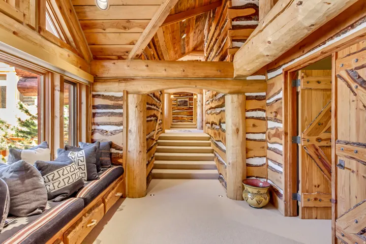 Log construction interior featuring exposed beams, a built-in window seat with storage drawers, a carpeted staircase, and rustic wood doors with iron hardware