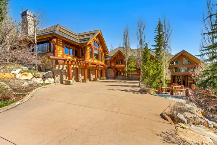 Rustic-modern log home featuring a stone chimney, extensive wood siding, multiple large windows, and a multi-car garage with overhead wood beams