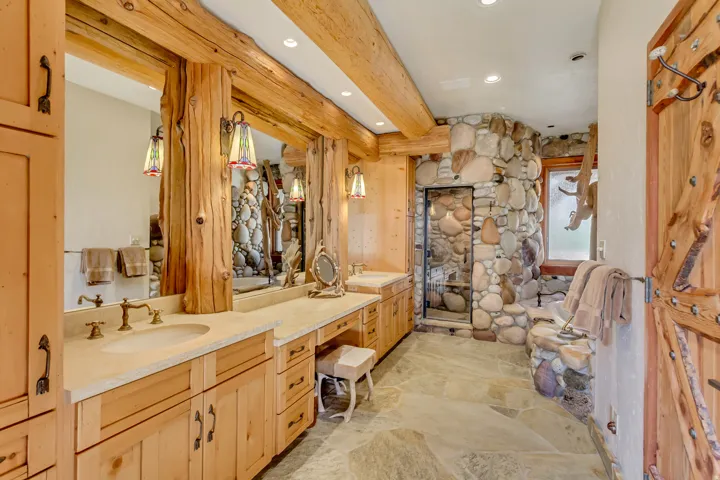 Bathroom featuring a stone-lined shower, natural wood beams, custom wood cabinetry, dual vanity with undermount sinks, and a stone slab floor