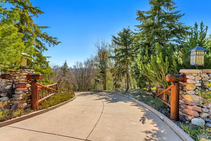 Gated entry drive featuring stone pillars, natural wood accents, and integrated lantern lighting, surrounded by mature evergreen trees and natural landscaping