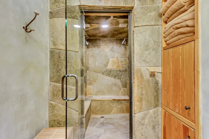 Walk-in shower featuring natural stone walls and flooring, a built-in bench, two showerheads, and recessed lighting