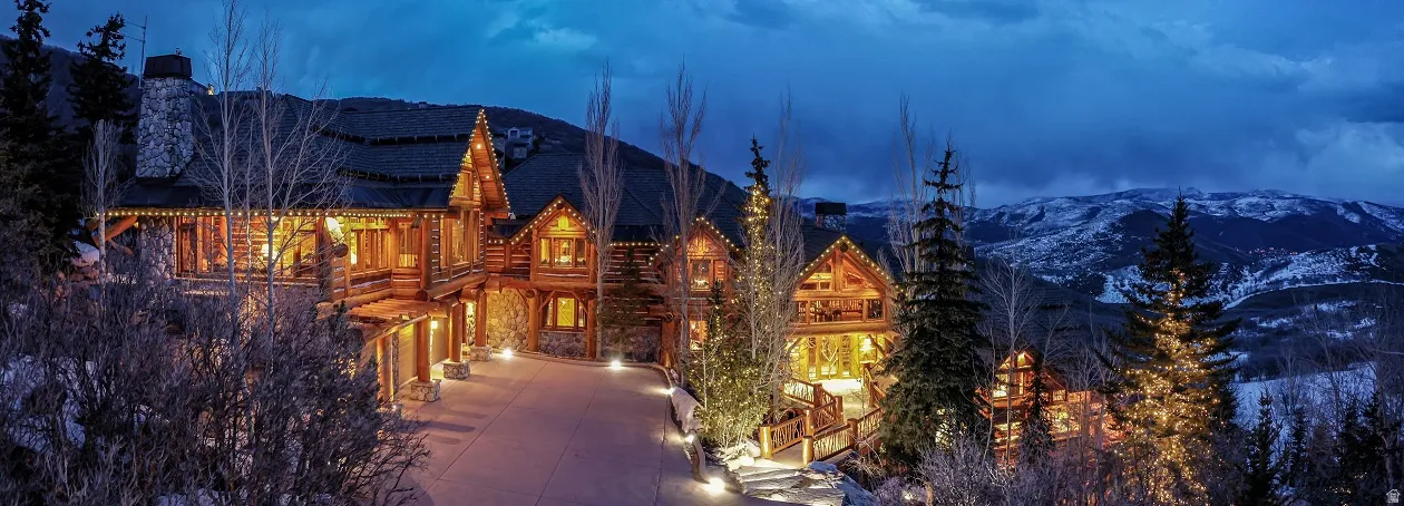Expansive multi-level log and stone residence featuring a stone chimney, extensive exterior lighting, a paved driveway, and multiple balconies