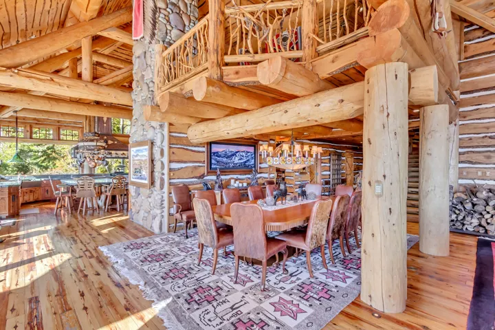 Expansive log cabin interior featuring a dining area with a chandelier, wood-finish flooring, exposed log beam ceilings, a stone column, and a kitchen with bar seating
