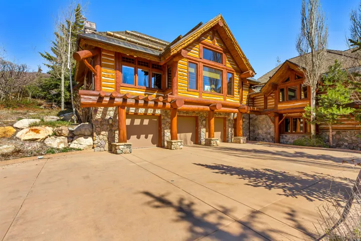 Rustic log home exterior featuring a multi-car garage, stone foundation, natural wood siding, and prominent windows, all situated on a paved driveway