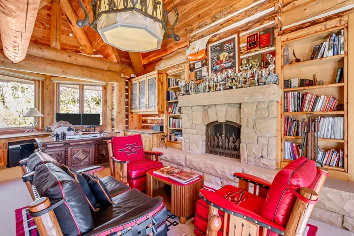 Open-concept room featuring exposed log beam construction, a stone-clad fireplace with an iron grate, built-in wooden shelving, and a large desk area with multiple windows
