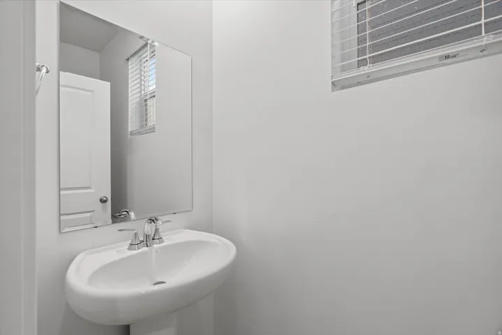 Pedestal sink with chrome faucet, large frameless mirror, and a window with blinds