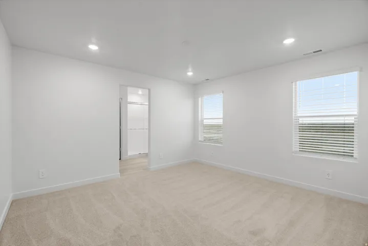 Spacious room featuring neutral carpeting, white walls, two windows with blinds, recessed lighting, and an entryway to an adjoining room with wood-finish flooring and a glass-enclosed shower