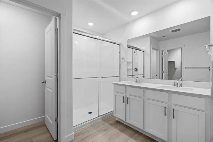 Bathroom featuring a dual vanity with white cabinetry and integrated sinks, a wide mirror, a glass-enclosed shower with white subway tile, and wood-finish flooring