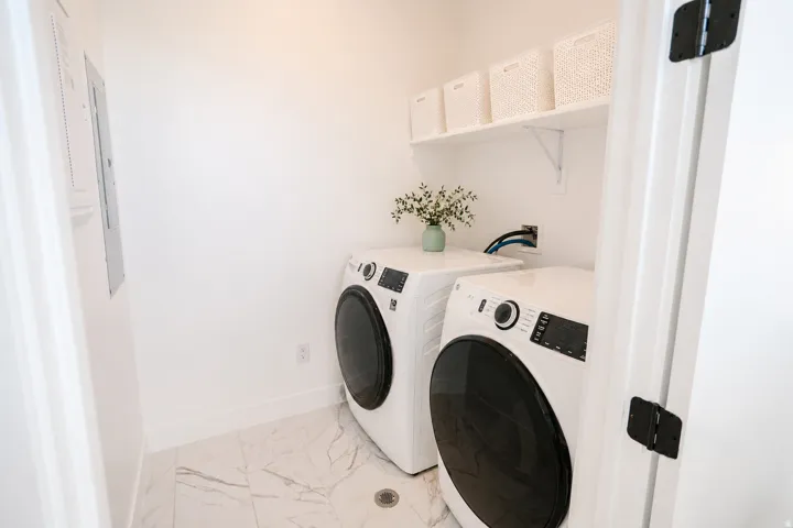 Dedicated laundry area featuring a white floating shelf, wall-mounted electrical panel, and white tile flooring with subtle veining