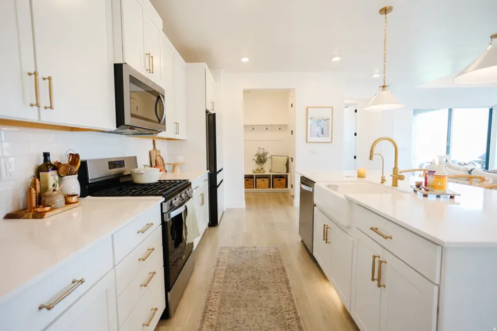 White cabinetry with gold-tone hardware, stainless steel appliances, white subway tile backsplash, light wood-finish flooring, and a farmhouse-style sink