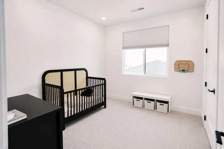 Bright nursery featuring a large window with cellular shade, neutral carpet flooring, black metal crib with natural wood accents, black dresser with ample storage, and built-in wall-mounted woven storage basket