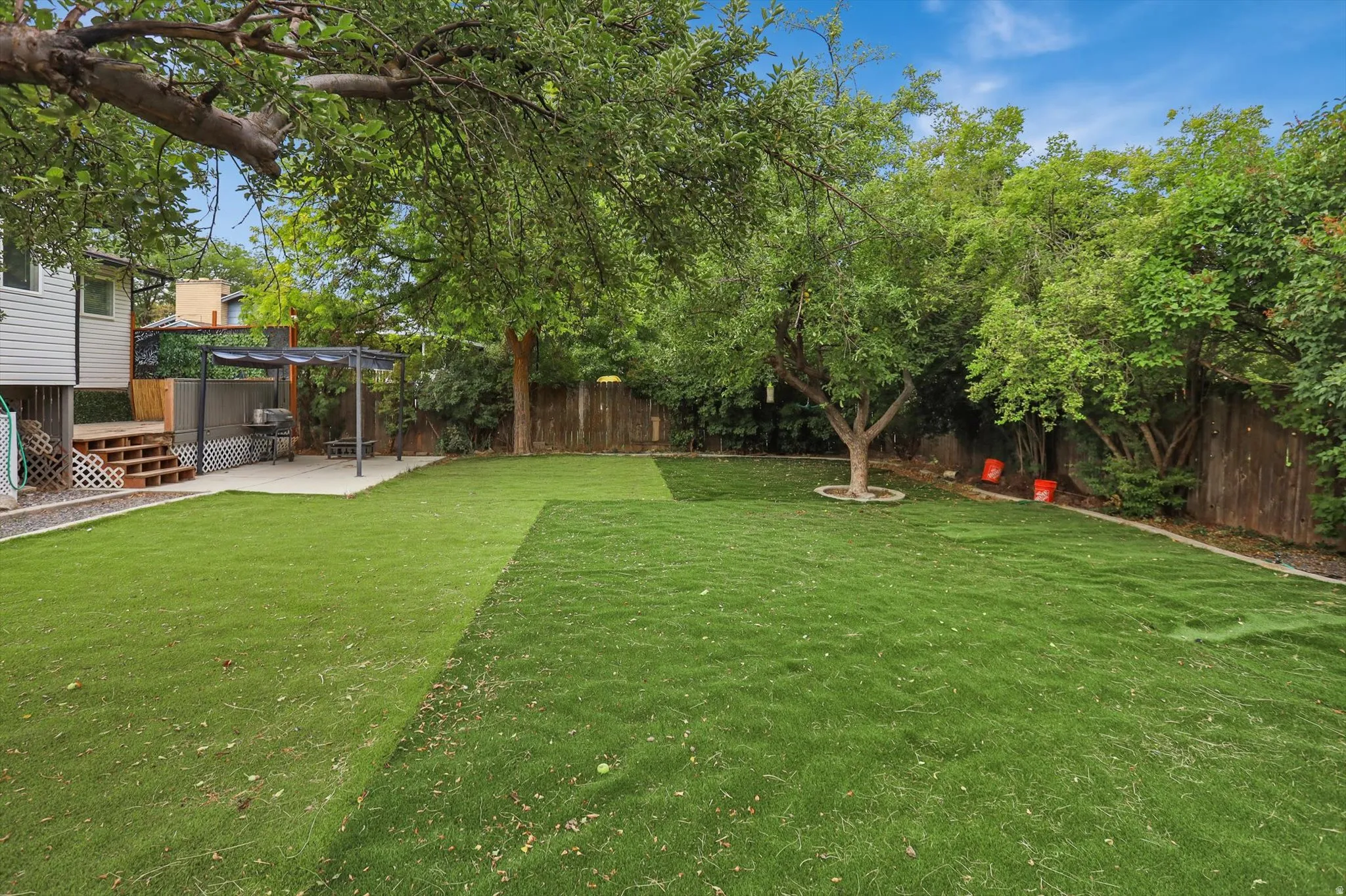Expansive green artificial lawn with mature shade trees, a concrete patio, and a built-in pergola