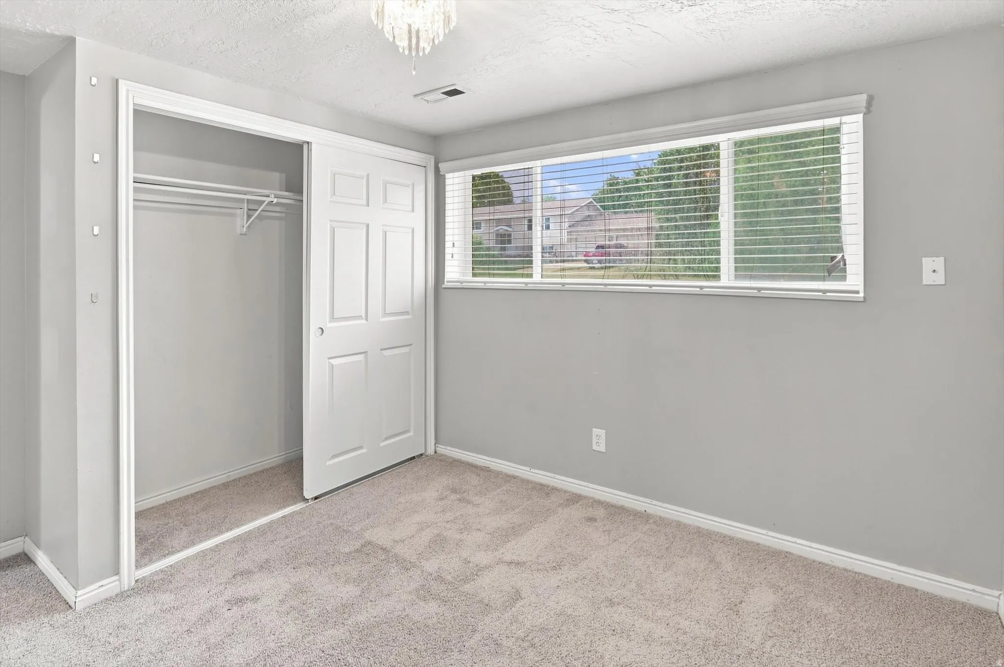 Carpeted room featuring a ceiling-mounted light fixture, sliding closet doors, a built-in closet organization system, and multiple windows with horizontal blinds