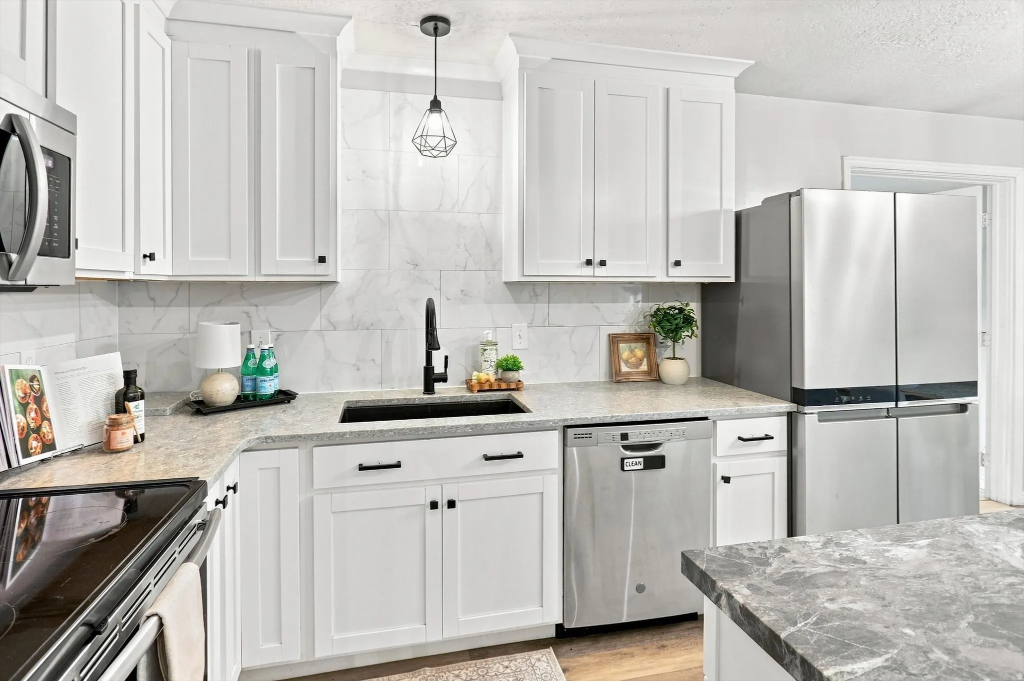 Kitchen featuring white shaker-style cabinetry with matte black hardware, light-toned countertops, full-height marble-look tile backsplash, stainless steel appliances, and a black sink with a matte black faucet